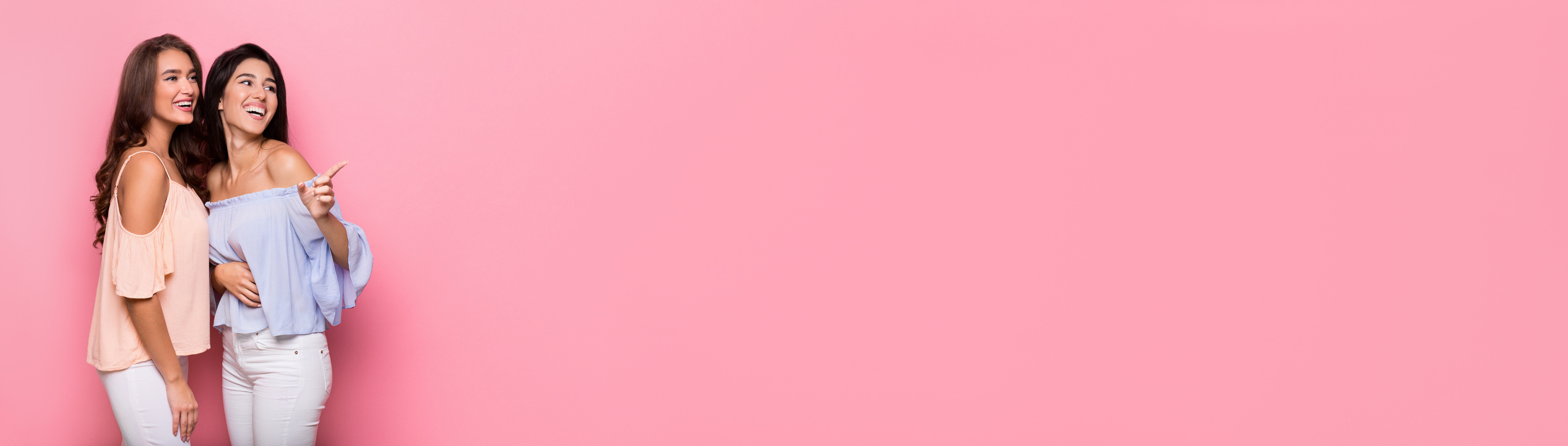 Two happy women point aside at pink background